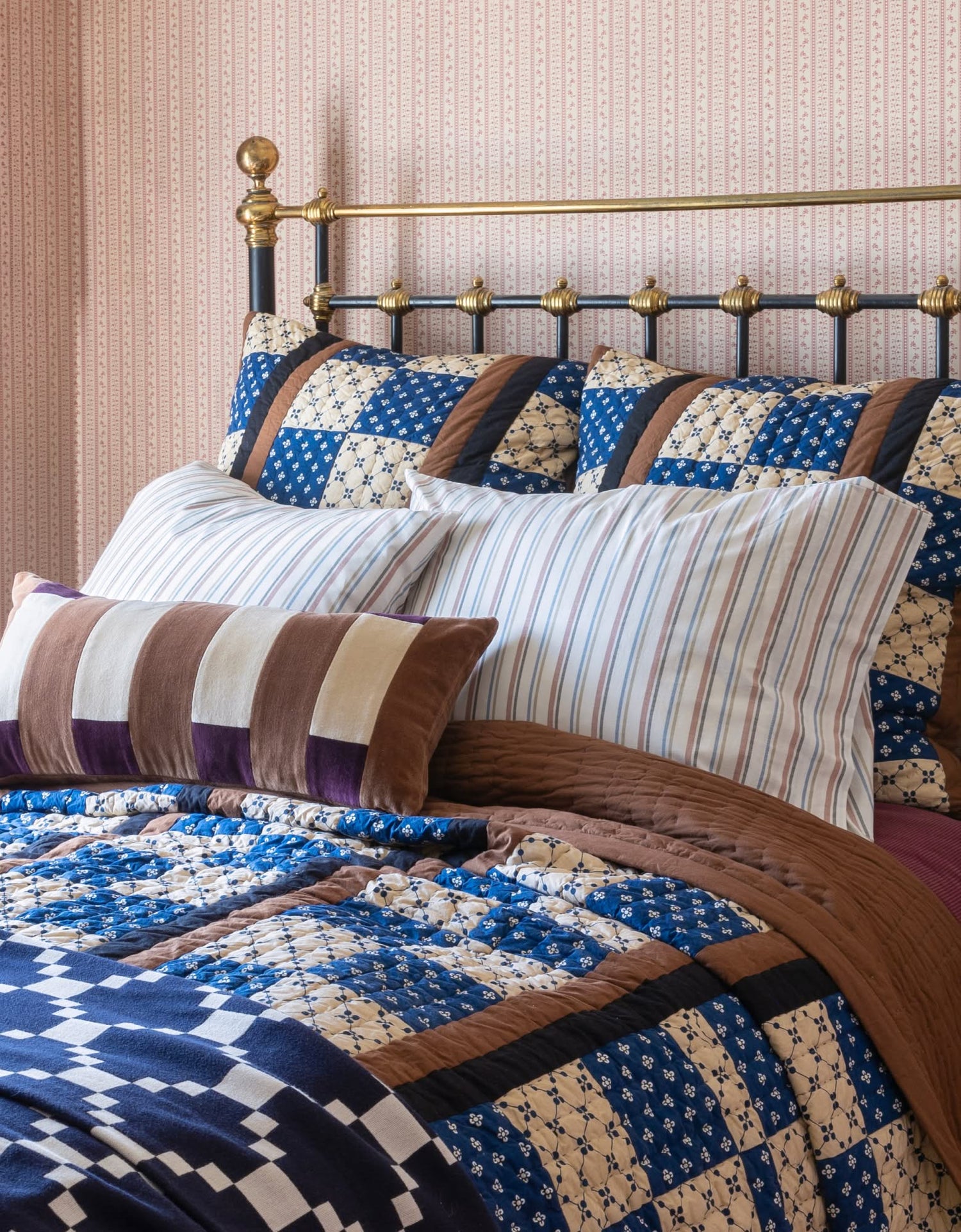 Angled view of patchwork quilt and layered bedding showing blue-and-cream patchwork, brown quilt, striped pillows on brass bed