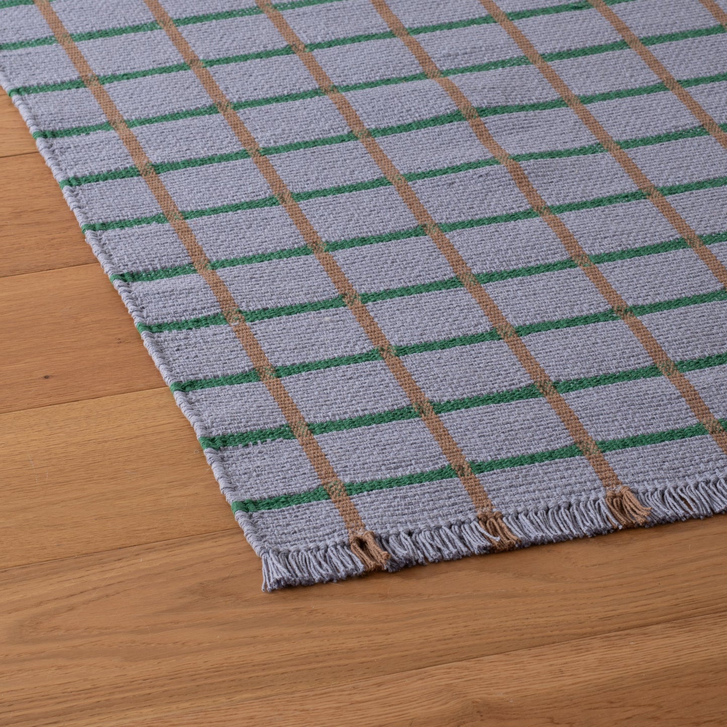 Angled close-up of Cotton Grid Flatweave Rug showing light-gray weave with green and tan grid stripes and fringe on wood floor