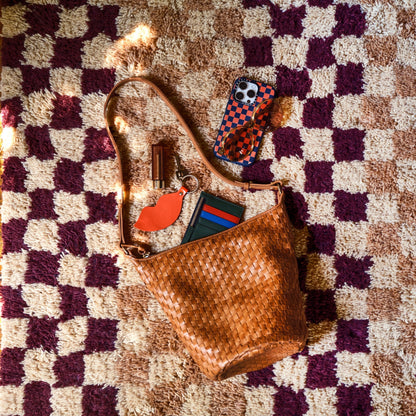 Top-down view of Schoolhouse x Clare V. Felix Shag Wool Rug in raisin and butterscotch checker pattern showcasing high-pile wool texture with a woven leather purse and phone