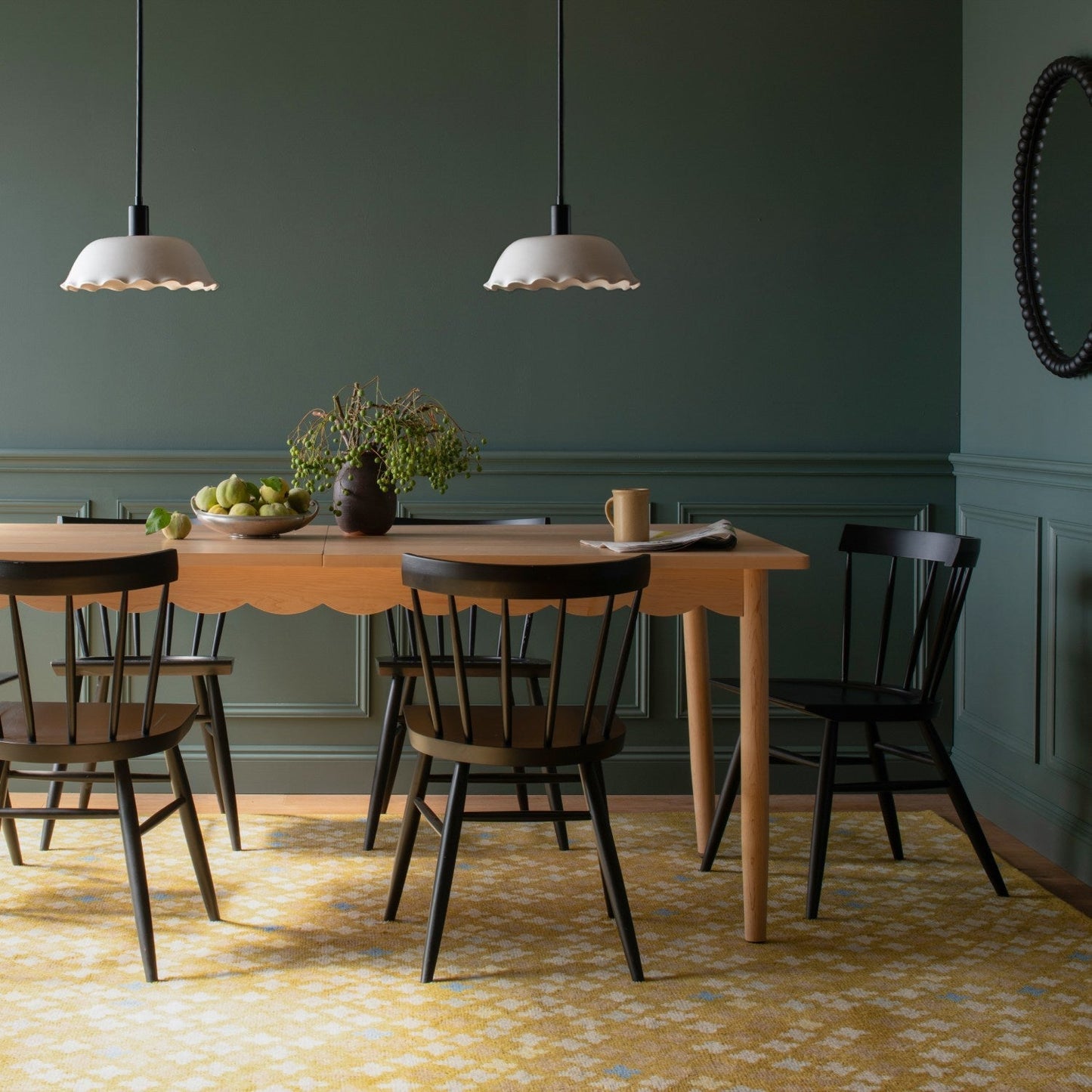 Bobbin mirror on a green paneled dining room wall above a scalloped edge wooden table with black chairs and pendant lights