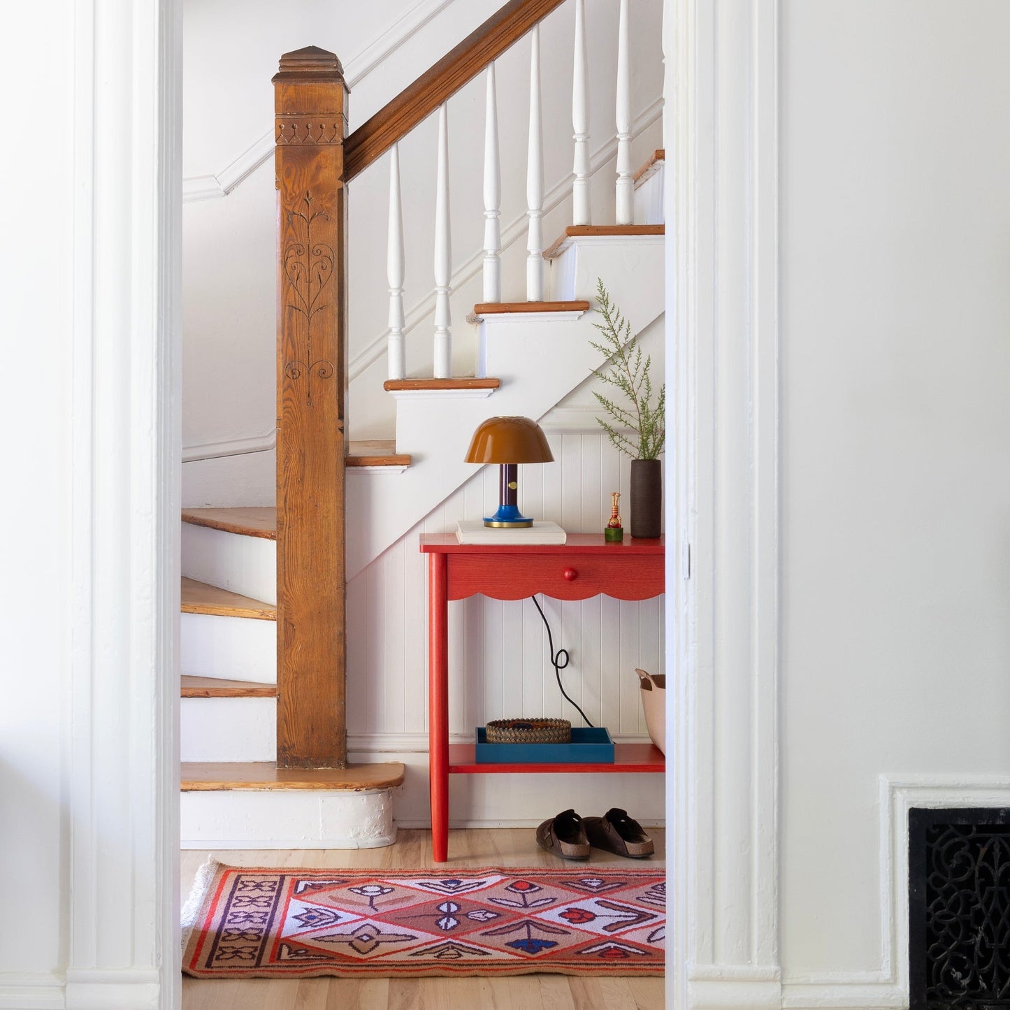 Linnea wool flatweave rug in an entryway beside a carved wood newel post, white staircase, and red side table with lamp