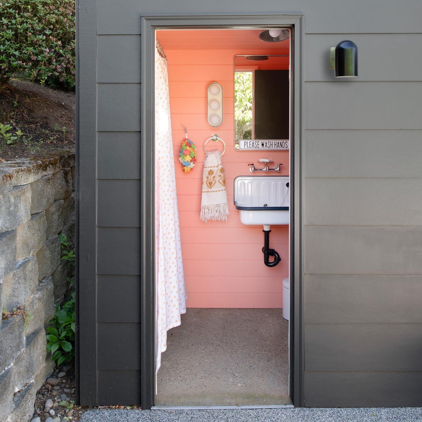 Front view of Allegheny 4.5-inch sconce in satin black, mounted on gray siding beside open doorway showing a pink-walled bathroom